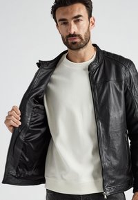 Mann, der eine schwarze Lederjacke aufmacht, um den weißen Pullover darunter zu zeigen, steht vor einem schlichten hellen Hintergrund.