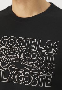 Černé tričko s bílým grafickým designem a opakujícím se textem "Lacoste" a ilustrací krokodýla. Vyrobeno z měkké bavlněné tkaniny.