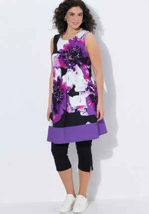 Frau mit lockigem Haar, die ein ärmelloses lila-schwarzes geblümtes Kleid über schwarzen Capri-Leggings und weißen Sneakers trägt, vor einer weißen Wand stehend.