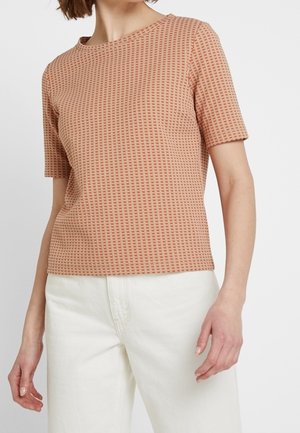 Frau trägt ein kurzärmeliges, strukturiertes, lachsfarbenes Oberteil und eine hoch taillierte weiße Hose, steht vor einem schlichten Hintergrund.