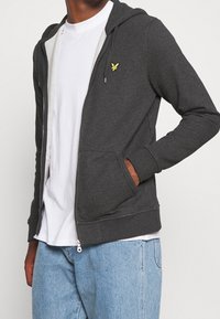 Persona con una felpa grigia scura non zipata con logo giallo, maglietta bianca e jeans azzurri chiari, mano nella tasca della felpa.