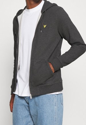 Persona con una felpa grigia scura non zipata con logo giallo, maglietta bianca e jeans azzurri chiari, mano nella tasca della felpa.