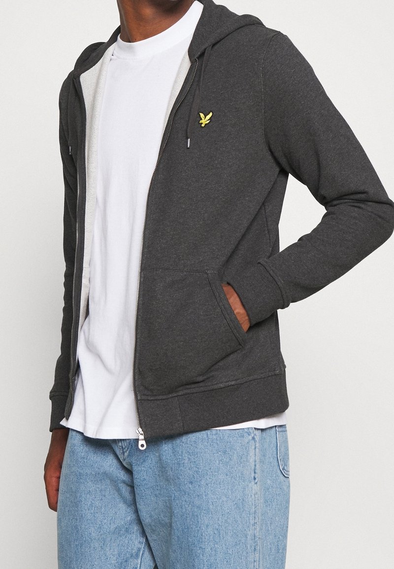 Persona con una felpa grigia scura non zipata con logo giallo, maglietta bianca e jeans azzurri chiari, mano nella tasca della felpa.