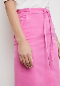 Vaaleanpunainen puuvillahame, jossa on sidottu vyötärö, sivutaskut ja valkoinen ommel. Sileä, hieman kiiltävä pinta.