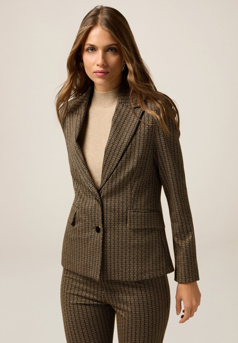 Blazer marron à motif avec une coupe ajustée, fermeture à un bouton et revers crantés. Porté sur un pull à col roulé beige.