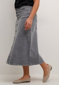 Siva denim midi krila z značkami spredaj, visokim pasom in razširjenim oblikovanjem, v kombinaciji s bež ploščatimi čevlji. Gladka tekstura.