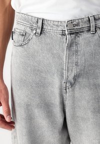 Jeans din denim gri cu o ușoară decolorare, având cinci buzunare, închidere frontală cu buton și cusături contrastante de-a lungul cusăturilor.