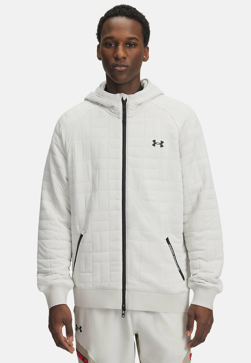 Haut à capuche blanc matelassé avec zip et accents noirs, doté d'une capuche, de poignets élastiques et de poches latérales. Logo visible sur la poitrine.