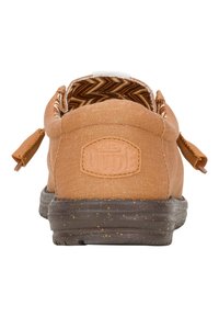 Zapatillas de lona color beige con una pestaña trasera, suela de goma marrón texturizada y forro con patrón de zigzag visible en el cuello. Logo "Hey Dude" en la parte posterior.