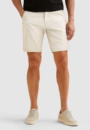 Mann trägt beige Shorts und passende beige Sneakers, steht vor einem einfarbigen hellgrauen Hintergrund.