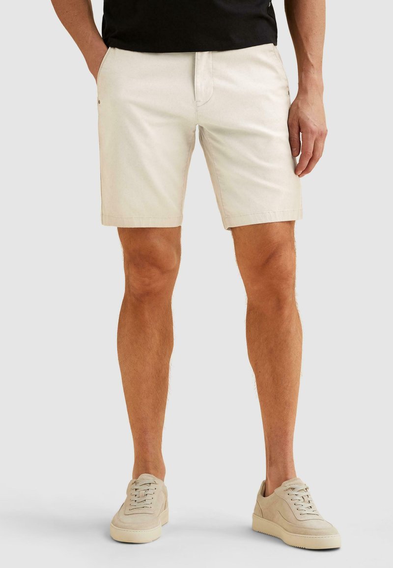 Homme portant un short beige et des baskets beige assorties, debout devant un fond gris clair uni.
