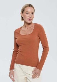 Haut à manches longues couleur rouille avec une texture côtelée et un encolure dégagée, forme ajustée et minimalisme dans le détail. Porté avec un pantalon de couleur claire.