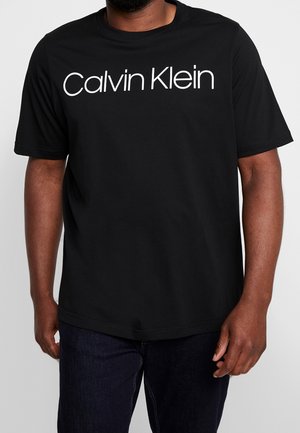 Mand iført en sort t-shirt med korte ærmer med hvid "Calvin Klein" tekst over brystet og mørkeblå jeans.
