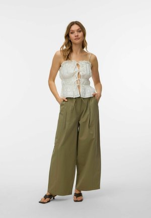 Femme debout, les mains dans les poches, portant un haut blanc sans manches avec liens à l'avant, un pantalon large et ample couleur olive et des talons ouverts noirs.