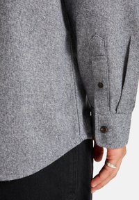 Gray textured shirt made of wool blend, featuring a buttoned cuff, with subtle color variation and a curved hem.