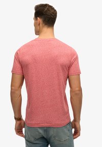 Camiseta de manga corta de color rojo jaspeado con escote redondo; textura suave y ajuste casual; vista desde atrás, mostrando los hombros y la parte superior de la espalda.