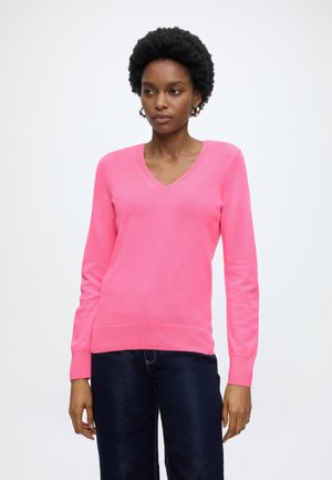 Vrouw draagt een felroze trui met V-hals en donkerblauwe spijkerbroek, staande tegen een eenvoudige witte achtergrond.