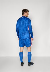 Tricou și pantaloni scurți sportivi de culoare albastră cu modele texturate, accente roșii pe margini și șosete albastre. Cizme de fotbal negre completează ținuta.
