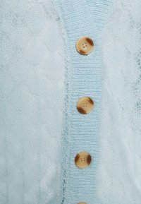 Jasnoniebieski kardigan o koronkowej fakturze z prążkowanym, dzianym kołnierzem. Z przodu znajduje się pięć dużych brązowych guzików, które pełnią funkcję zapięcia.