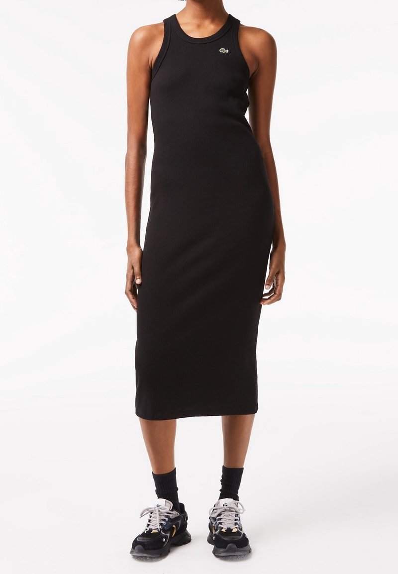 Femme portant une robe midi noire sans manches et des chaussettes noires avec des baskets noires et blanches, debout devant un fond blanc.