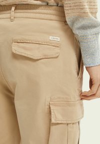 Beige cargobukser med klaplommer på bagsiden og siden, båret med en flerfarvet striktrøje synlig ved håndleddet.