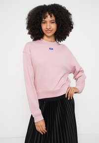 Sudadera rosa claro con puños y dobladillo acanalados, que presenta un pequeño parche de logo azul en la parte delantera. Combinada con una falda negra plisada.