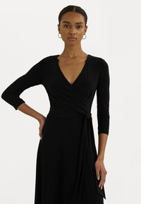 Black wrap dress with a deep V-neck, long sleeves, and a tied accent at the waist. Smooth, stretchy fabric with a flowing silhouette.