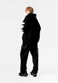 Sudadera negra de gran tamaño con rayas gráficas blancas en las mangas, combinada con pantalones de chándal negros que presentan un detalle de cadena y calzado negro voluminoso.