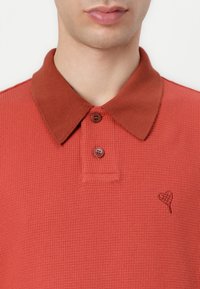 Rustfarget polo skjorte med teksturert stoff, med brun krage, to knapper og en hjerteformet broderidetalj.
