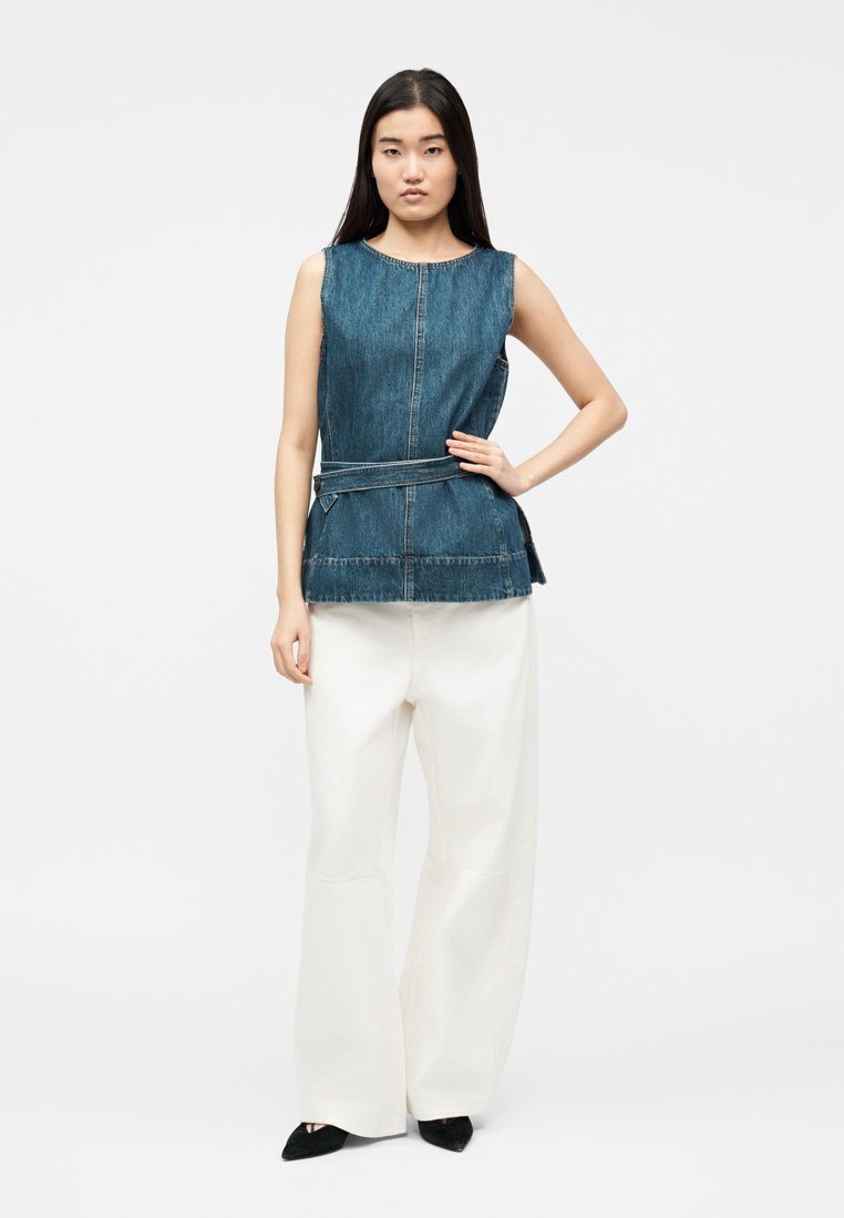 Femme debout, main sur la hanche, portant un haut en denim sans manches avec ceinture et un pantalon blanc ample, sur fond blanc.