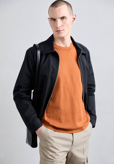 Veste zippée noire avec col, associée à un pull orange à col rond et un pantalon beige. Comprend des poches classiques et un design élégant.