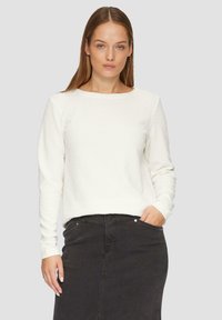 Pull manches longues texturé blanc avec un col rond, associé à une jupe en denim foncé. Le tissu semble légèrement surélevé, ajoutant de la dimension.