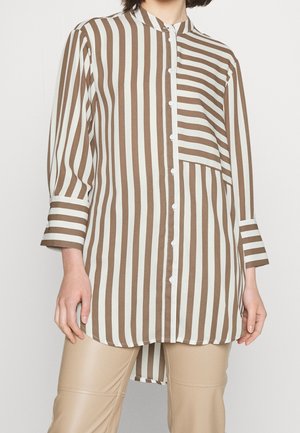 Person trägt ein braun-weiß vertikal gestreiftes Hemd mit Knöpfen und langen Ärmeln sowie beige Hosen.