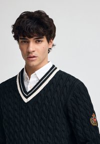 Svart kabelstickad tröja med en vit och svart randig V-hals. Har en logotypapplikation på vänster ärm. Strukturerad tyg.