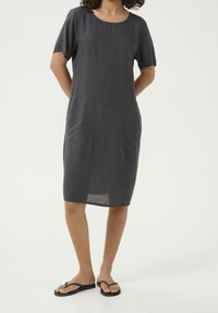 Harmaa pellavamekko lyhyillä hihoilla, pyöreällä kaula-aukolla ja sivutaskuilla. Yksinkertainen pinta, polvipituinen, yhdistettynä yksinkertaisiin mustiin läpsyköihin.