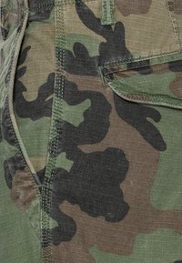 Zielony, brązowy i czarny materiał w kamuflażu z widocznymi szwami oraz częściowo otwartą kieszenią klapową.