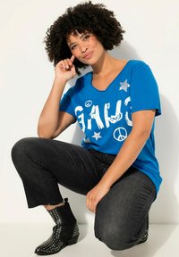 Camiseta de manga corta azul con texto en blanco en negrita y detalles de estrellas plateadas. Combinada con jeans de mezclilla negros y botas de tobillo negras con tachuelas.