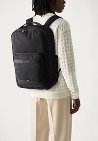 Black backpack made of durable fabric, featuring a front pocket, padded straps, and a subtle logo detail. Smooth texture with minimalistic design.