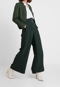 Tmavě zelený blazer s texturovaným povrchem, kombinovaný s vysokými, širokými zelenými kalhotami s předními puky a zlatými knoflíky. Pod ním bílá blůza a krémově zbarvené kotníkové boty.