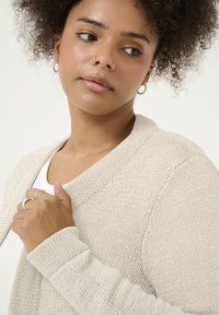 Cardigan en tricot beige avec un col en V, des poignets côtelés et une finition texturée. Modèle le tenant légèrement au niveau de la poitrine, portant un haut blanc en dessous.