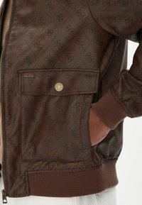 Giubbotto bomber in tessuto trapuntato marrone con motivo logo in rilievo, dotato di chiusura con zip e polsini a coste. Include una tasca frontale con bottone a pressione.