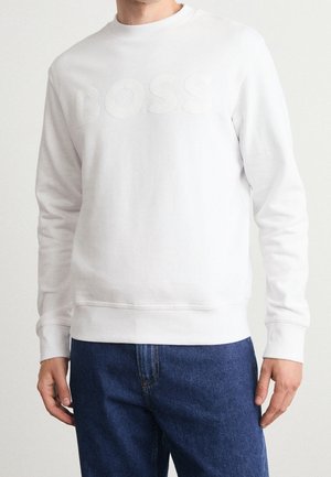 Homme portant un sweat-shirt blanc avec le logo "BOSS" embossé et un jean bleu, debout devant un fond blanc uni.