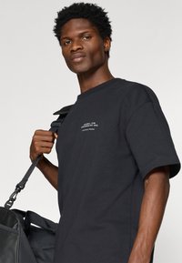 Melns kokvilnas T-krekls ar apļveida kakla izgriezumu un īsām piedurknēm, blakus melnai sporta somai ar siksnas detaļām.