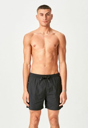 Fit young man standing facing forward, wearing black drawstring shorts, no shirt, neutral expression, plain light background.