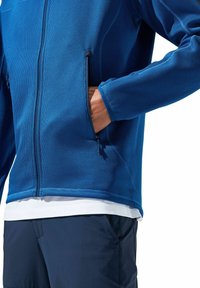 Blaue Zip-Jacke mit strukturiertem Oberflächen, ausgestattet mit zwei Seitentaschen und kontrastierenden Einsätzen, kombiniert mit dunklen Hosen und einem weißen Unterhemd.