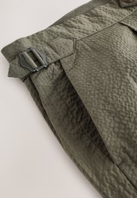 Tessuto verde oliva con finitura liscia e trama, caratterizzato da una fibbia in metallo nero e cuciture decorative lungo la vita e le pieghe.