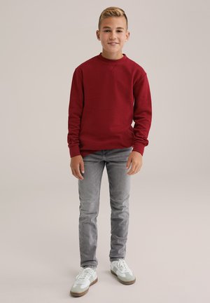 Dreng står oprejst, iført en rød sweater, grå jeans og hvide sneakers, smilende mod en lys, ensfarvet baggrund.