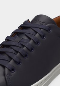 Αθλητικό παπούτσι από δέρμα ναυτικού μπλε χρώματος με κορδόνια στο ίδιο χρώμα, με ανάγλυφο λογότυπο στη γλώσσα και λευκή ελαστική σόλα, σε κοντινή λήψη πάνω σε ανοιχτό φόντο.