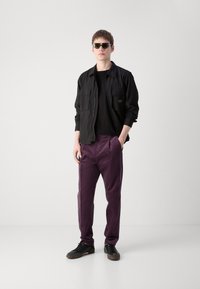 Veste noire avec deux poches sur la poitrine, t-shirt noir, pantalon violet et baskets noires. Les accessoires incluent des lunettes de soleil et une bague.