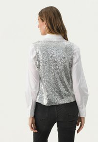 Sølvpalietvest med en tætsiddende pasform, båret over en hvid skjorte med krave, kombineret med sorte jeans, der viser en glitrende tekstur.